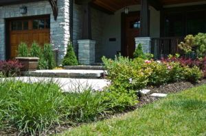 A stone walkway leads to the entrance of a house with a wooden garage door, surrounded by shrubs and mulched garden beds.