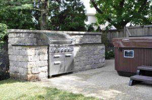 An outdoor kitchen featuring a stone grill island set on a patio next to a hot tub.