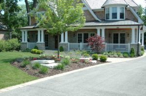 A multi-story house with a wrap-around porch, stone facade, and landscaped front yard featuring shrubs and a central tree.