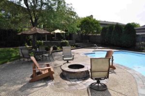 A stone fire pit sits on a patio next to a swimming pool, surrounded by various patio chairs and umbrellas in a backyard.