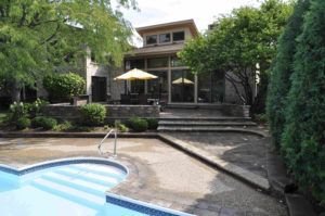A backyard patio features a swimming pool in the foreground, leading to stone steps, patio furniture, and a house exterior.