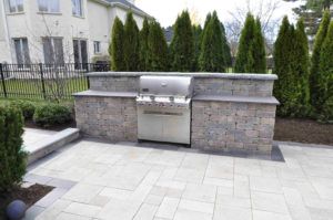 A built-in stainless steel grill set into a stone block island on a paved patio in a residential backyard.