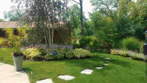 A stone stepping stone path winds through a green lawn toward a garden bed with shrubs, purple flowers, and trees.