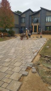 A worker pushes a wheelbarrow on a newly laid light-colored stone paver driveway in front of a modern two-story home.