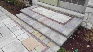 Two gray stone steps with a red brick border lead up to a doorway next to a paved walkway and a garden bed.