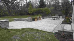 A stone patio with a curved seating wall, pillar lights, and outdoor dining furniture set in a grassy backyard.