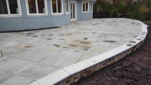 A newly installed gray stone patio with a curved retaining wall extending from the back of a house.