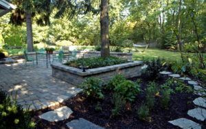 A stone patio with a raised planter bed, trees, and a flagstone path in a lush, green backyard setting.