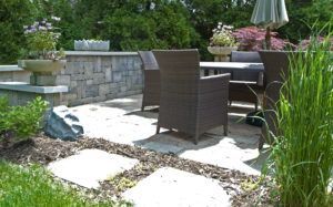 Patio dining area with wicker chairs and a stone wall, surrounded by garden beds, planters, and a stepping stone path.