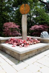 A vertical stone monument topped with a textured, circular orange sculpture in a garden bed with red flowers.