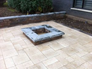 A square stone fire pit sits in the center of a paved stone patio near a retaining wall and the corner of a house.