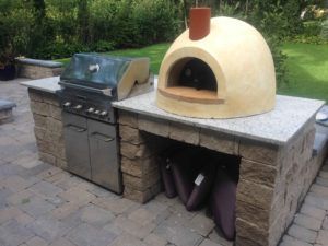 An outdoor kitchen on a stone patio featuring a stainless steel grill next to a beige, dome-shaped pizza oven.