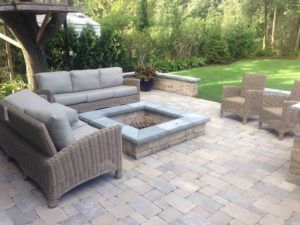 Patio area with wicker sofa and armchairs arranged around a square stone fire pit on a paver ground, surrounded by trees.