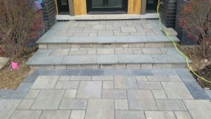 A stone paver walkway leads to a two-step concrete staircase in front of a house entrance.
