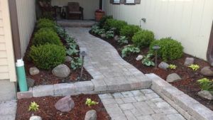 A stone paver walkway leads to a small porch between two beige house walls, flanked by manicured shrubs, mulch, and rocks.