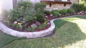 A curved stone garden border edging a mulched bed with shrubs, flowers, and stones against a house foundation.