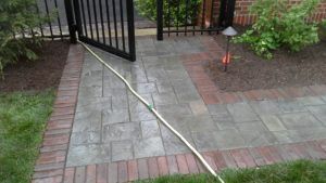A stone walkway with a red brick border leads to a black metal gate, with a white garden hose stretched across the path.