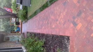 A red brick walkway framed by green grass and a garden bed, leading toward a house exterior with a sliding door.