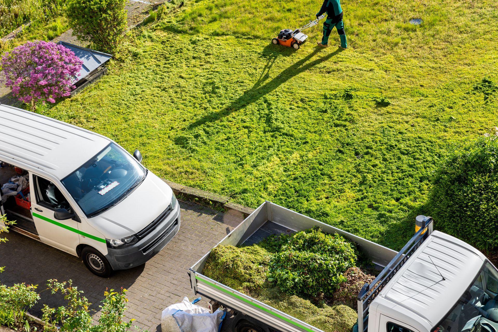 Landscape maintenance services crew mowing residential lawn with work trucks parked outside.
