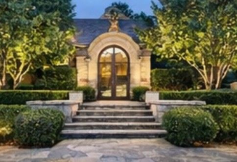 Stone building with arched doorway and steps, framed by hedges and trees, lit at dusk. An arched glass doorway leads into a stone-facade building, framed by symmetrical manicured bushes and stone steps.