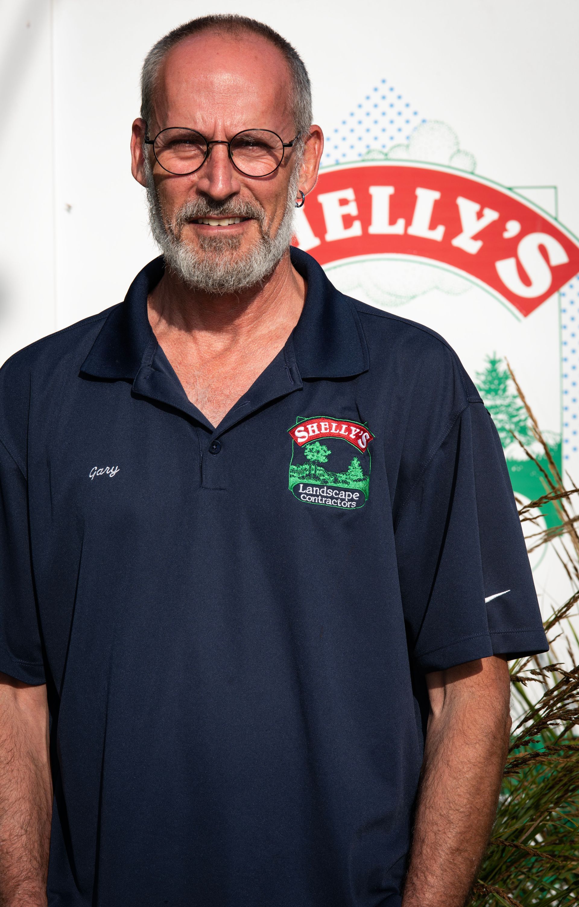 Gary — Northbrook, IL — Shelly’s Landscape A person with a beard and glasses wears a navy polo shirt with a logo, standing in front of a Kelly's brand sign.
