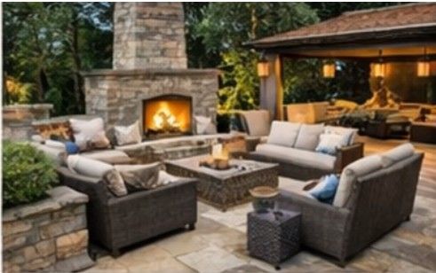 Luxury outdoor living space North Shore IL, natural stone patio, outdoor fireplace, outdoor kitchen, evening lighting, upscale backyard design A cozy outdoor patio with wicker seating arranged around a stone fireplace and fire pit under a covered structure.