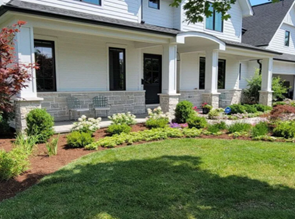 White two-story suburban home with a stone-based front porch, black window frames, and a landscaped yard with green shrubs.