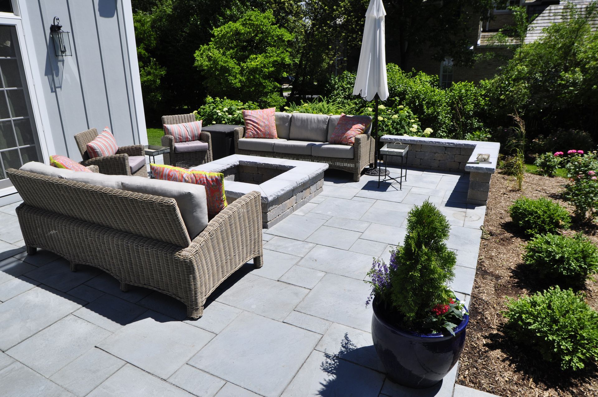 A light gray stone patio featuring a fire pit, wicker outdoor furniture, and a blue planter set against a garden backdrop.
