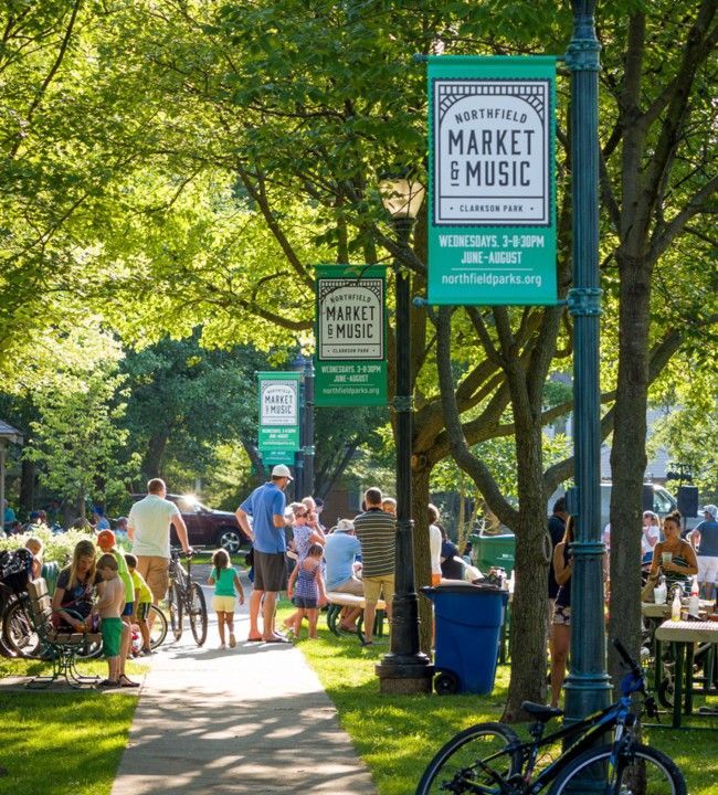Market and Music event in a park, people gathered near benches, banners on lamp posts. Green event banners hang from lampposts over a leafy park path filled with people walking, cycling, and sitting at tables.