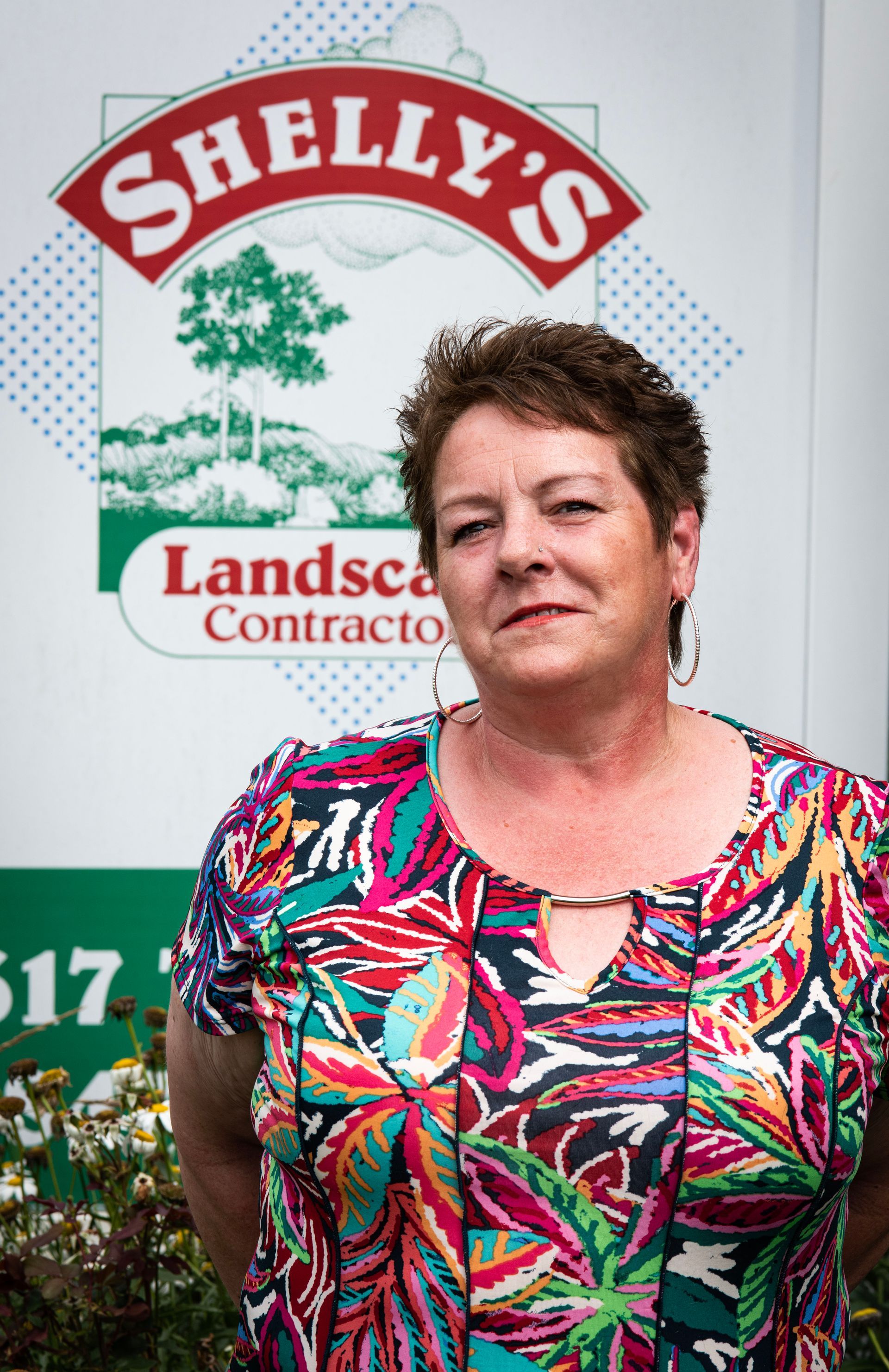 Patti — Northbrook, IL — Shelly’s Landscape A person stands in front of a branded sign for