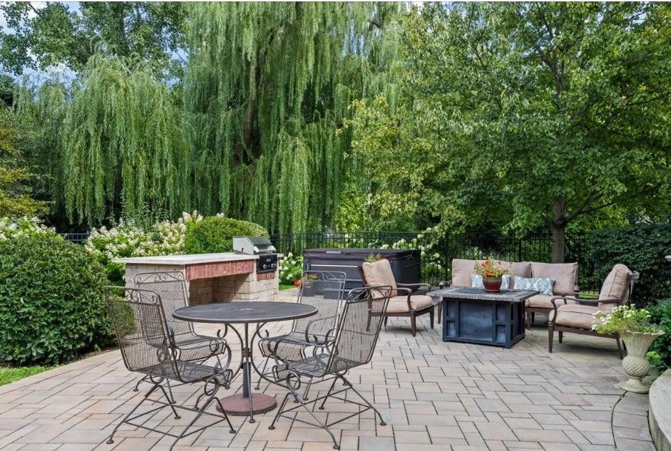 Two-part image: Well-manicured green lawns with trees, bushes, and buildings on a sunny day. A patio with an outdoor kitchen, a round dining table, and lounge seating surrounded by trees and lush greenery.