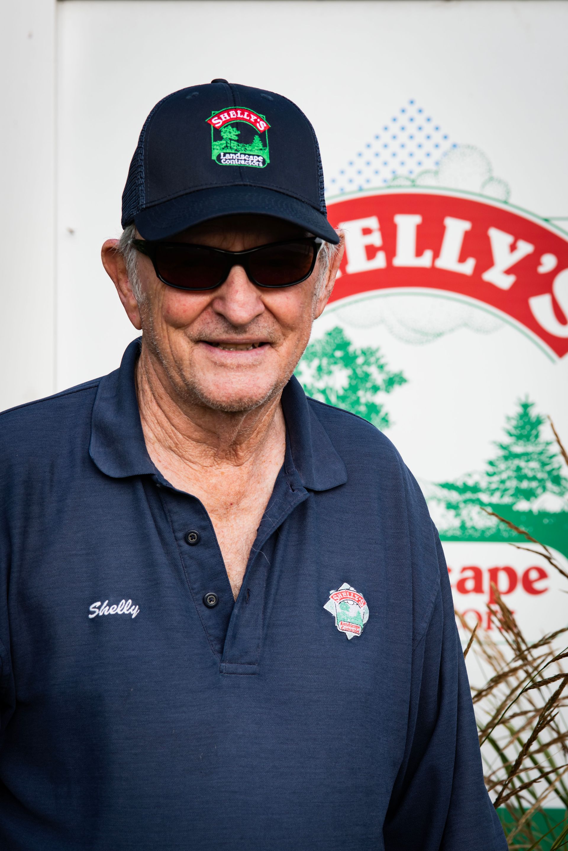 Shelly — Northbrook, IL — Shelly’s Landscape An older person wearing a dark blue polo shirt and a matching baseball cap stands in front of a Kelly’s sign.
