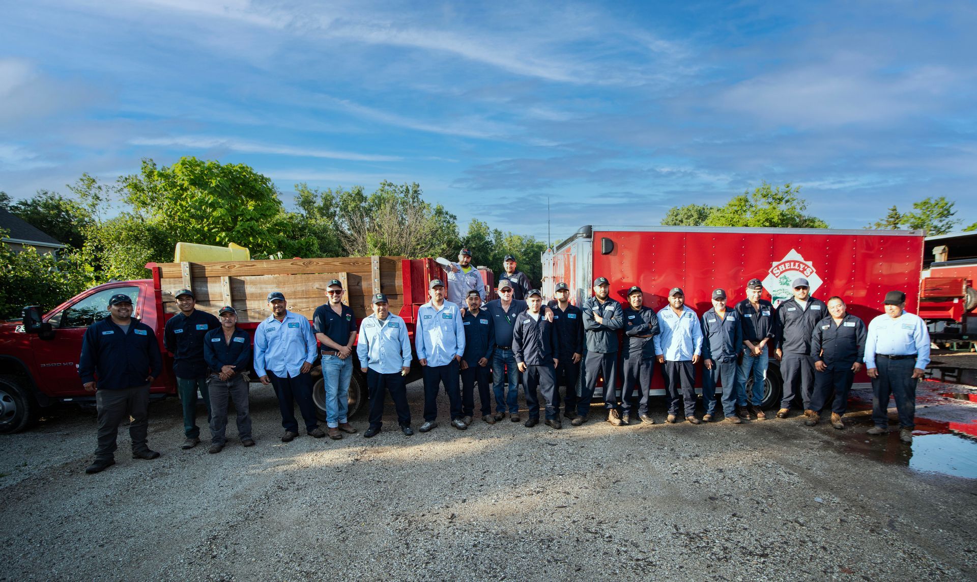 Team Member — Northbrook, IL — Shelly’s Landscape A group of employees in work uniforms standing in front of two large red trailers parked on a gravel lot.