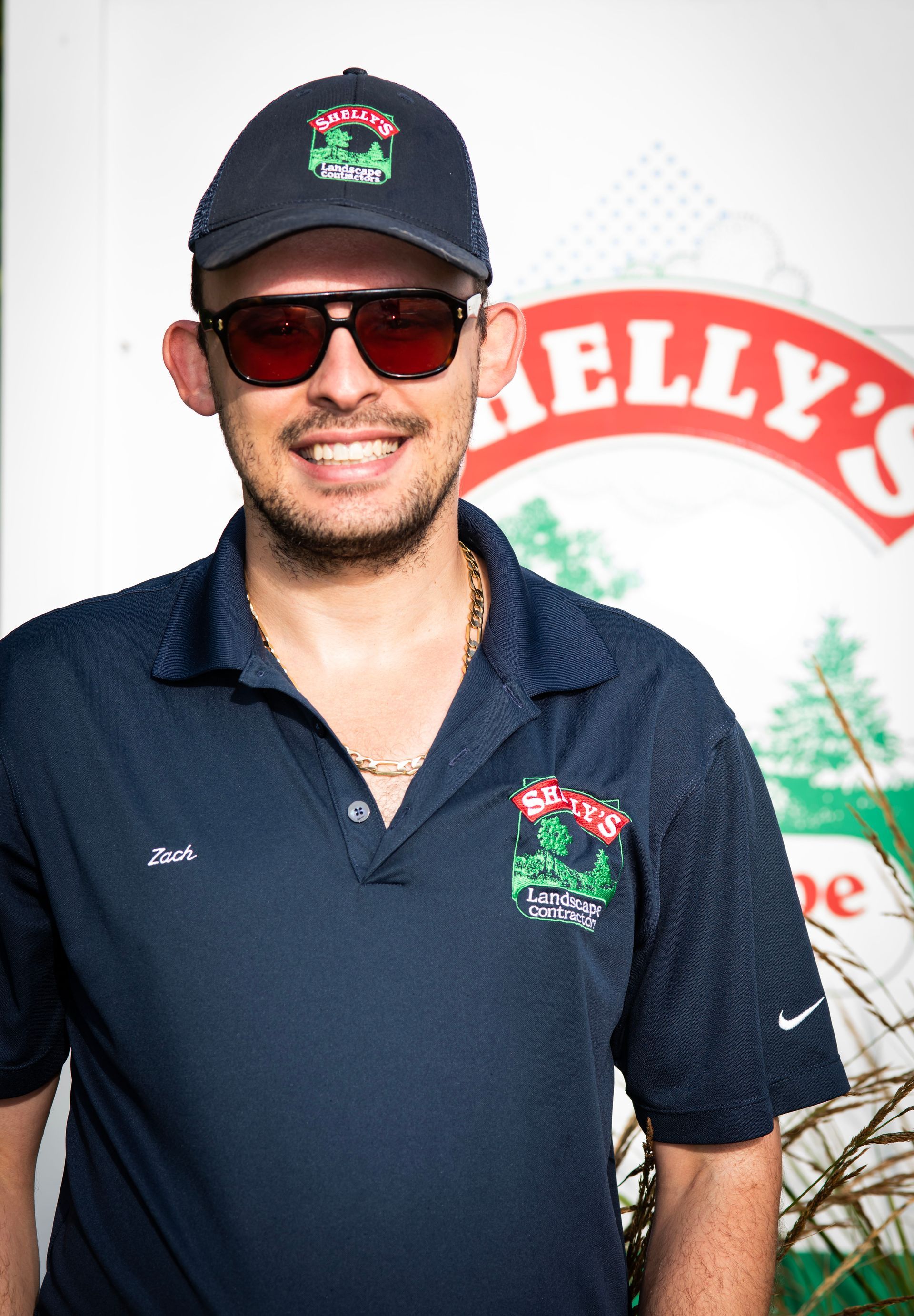 Zach — Northbrook, IL — Shelly’s Landscape A smiling individual wearing a dark blue polo shirt, matching baseball cap, and sunglasses, standing before a branded sign.
