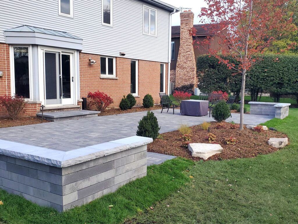 A backyard patio with a stone wall, paved seating area, landscaping, and a house exterior in the background.