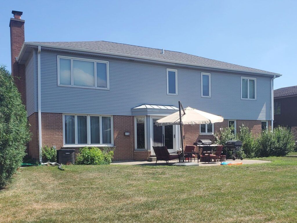 A two-story house with grey siding and a brick lower level, featuring a back patio with patio furniture and an umbrella.