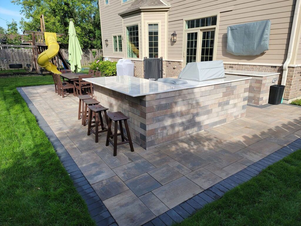 An outdoor patio with a stone bar, grill, and stools, featuring a dining area and a playground set in the background.