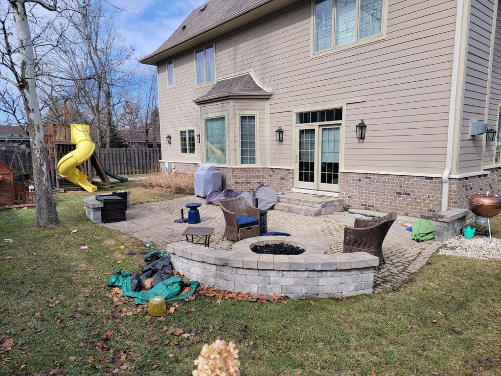 A tan house with a brick-paved patio, outdoor furniture, a fire pit, and a yellow slide in the background.