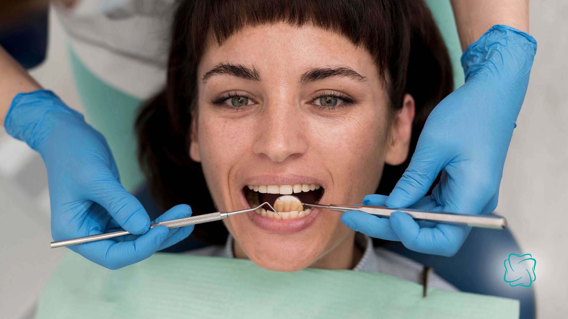 Woman at the dentist, mouth open, being examined with tools by someone wearing blue gloves.