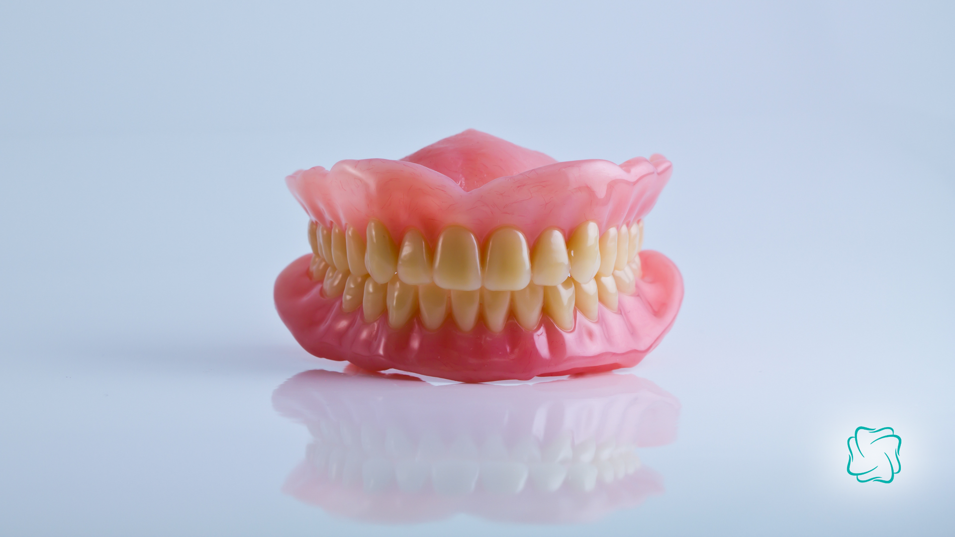 Full set of dentures, pink base with rows of artificial teeth, on a reflective white surface.