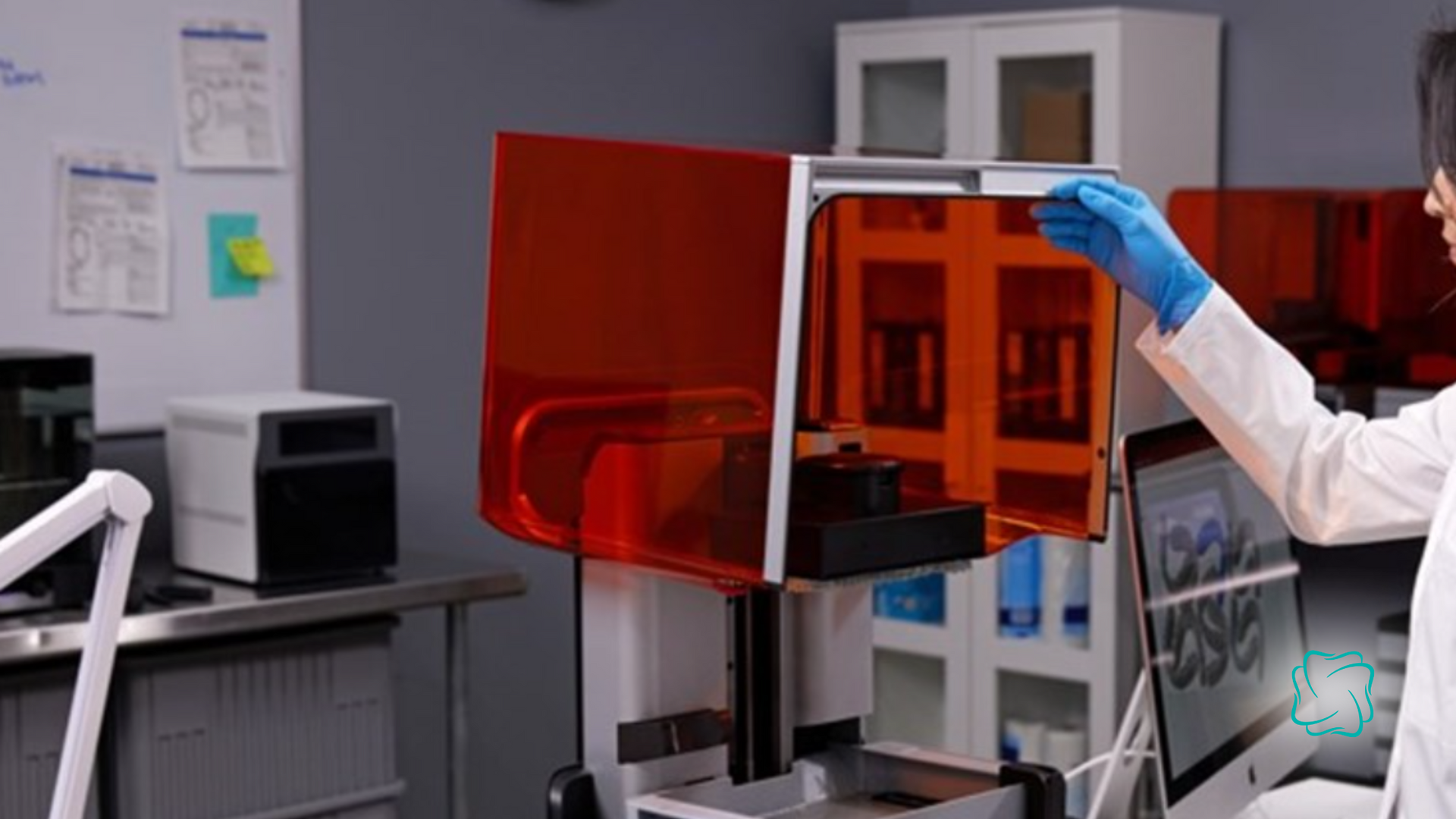 Person in lab coat opens a red 3D printer in a laboratory. Computer screen shows graphics.