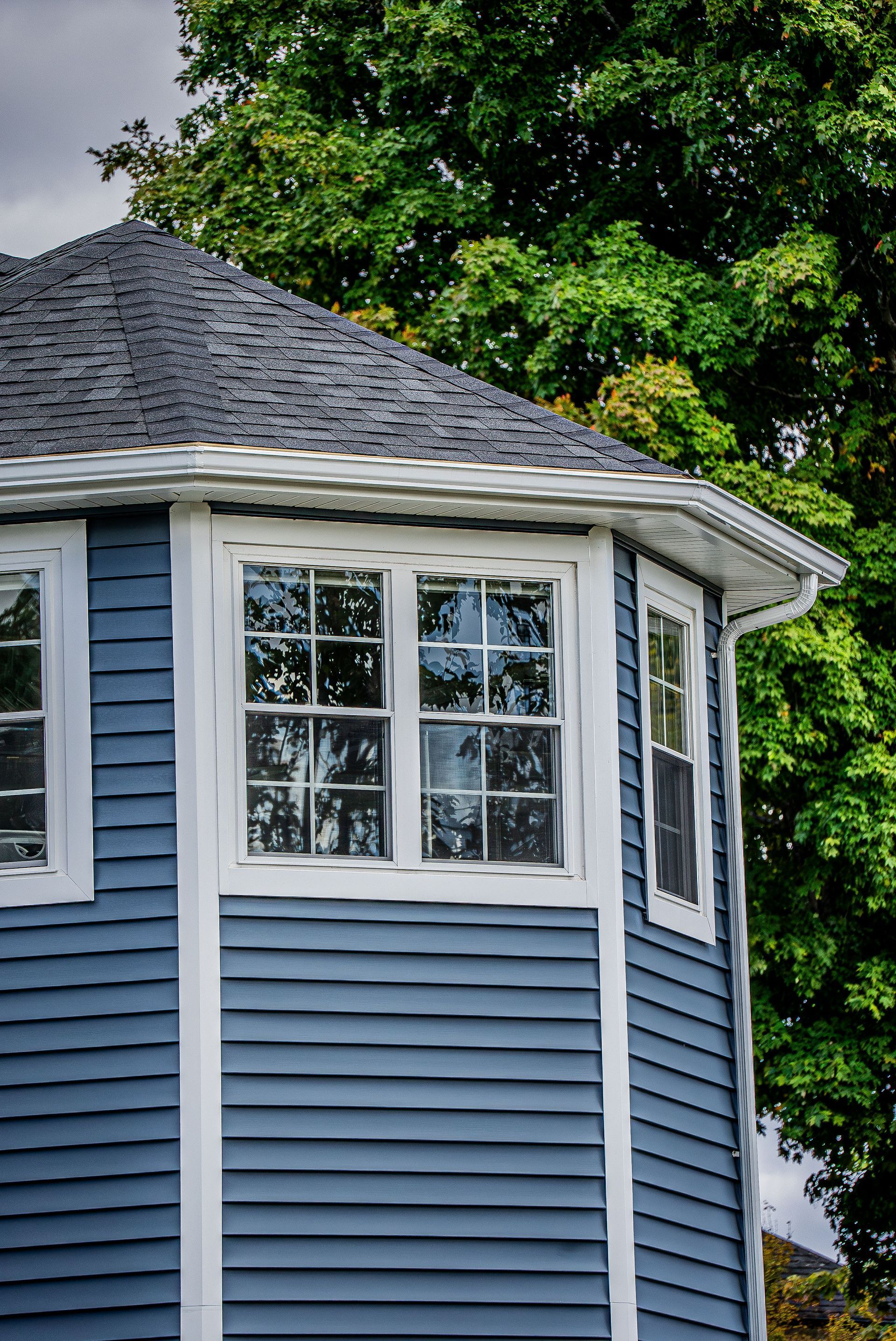 Blue house with white trim, bay window, and dark roof, with green tree in background.
