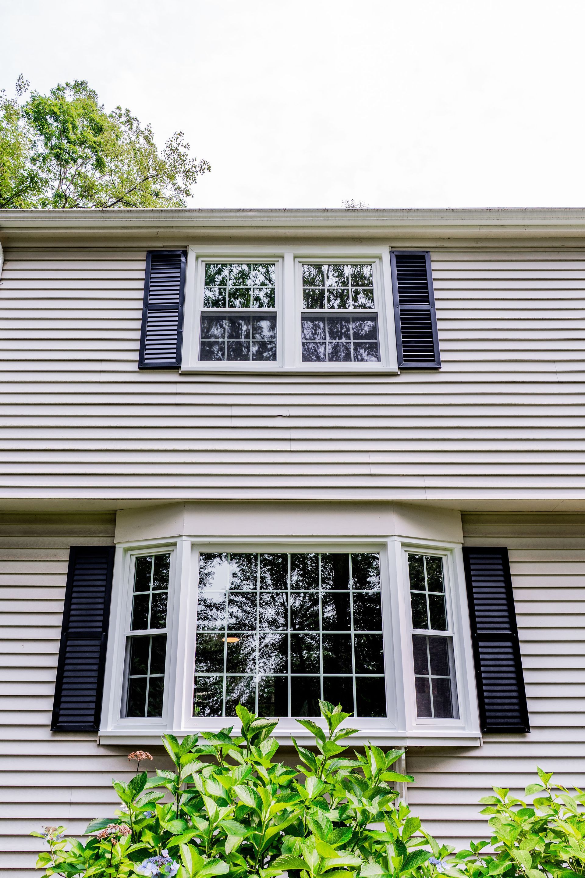Two-story house with white siding, black shutters, and windows. Lush green bushes are in the foreground.