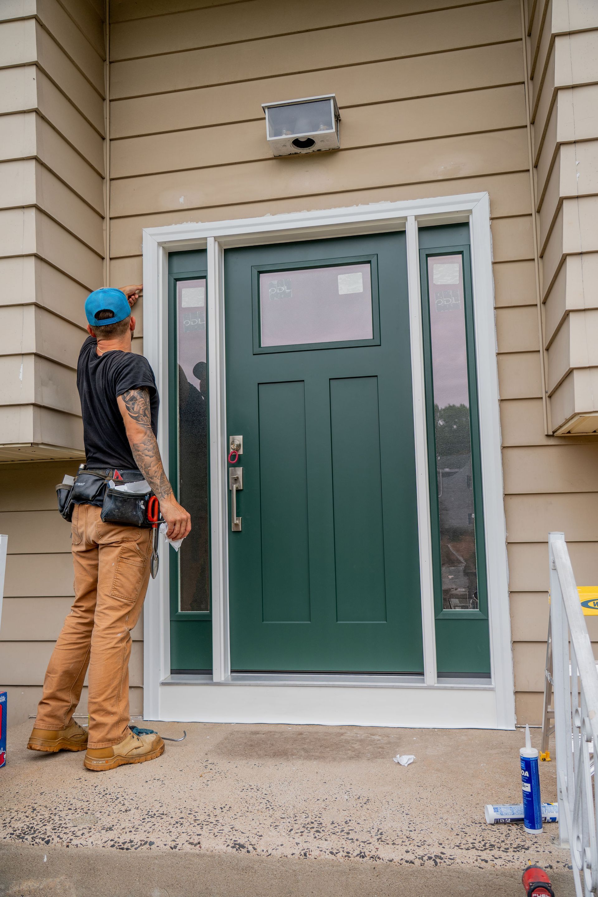 Carpenter installing white trim around a green front door. Man is tattooed, wearing work clothes, on a porch.