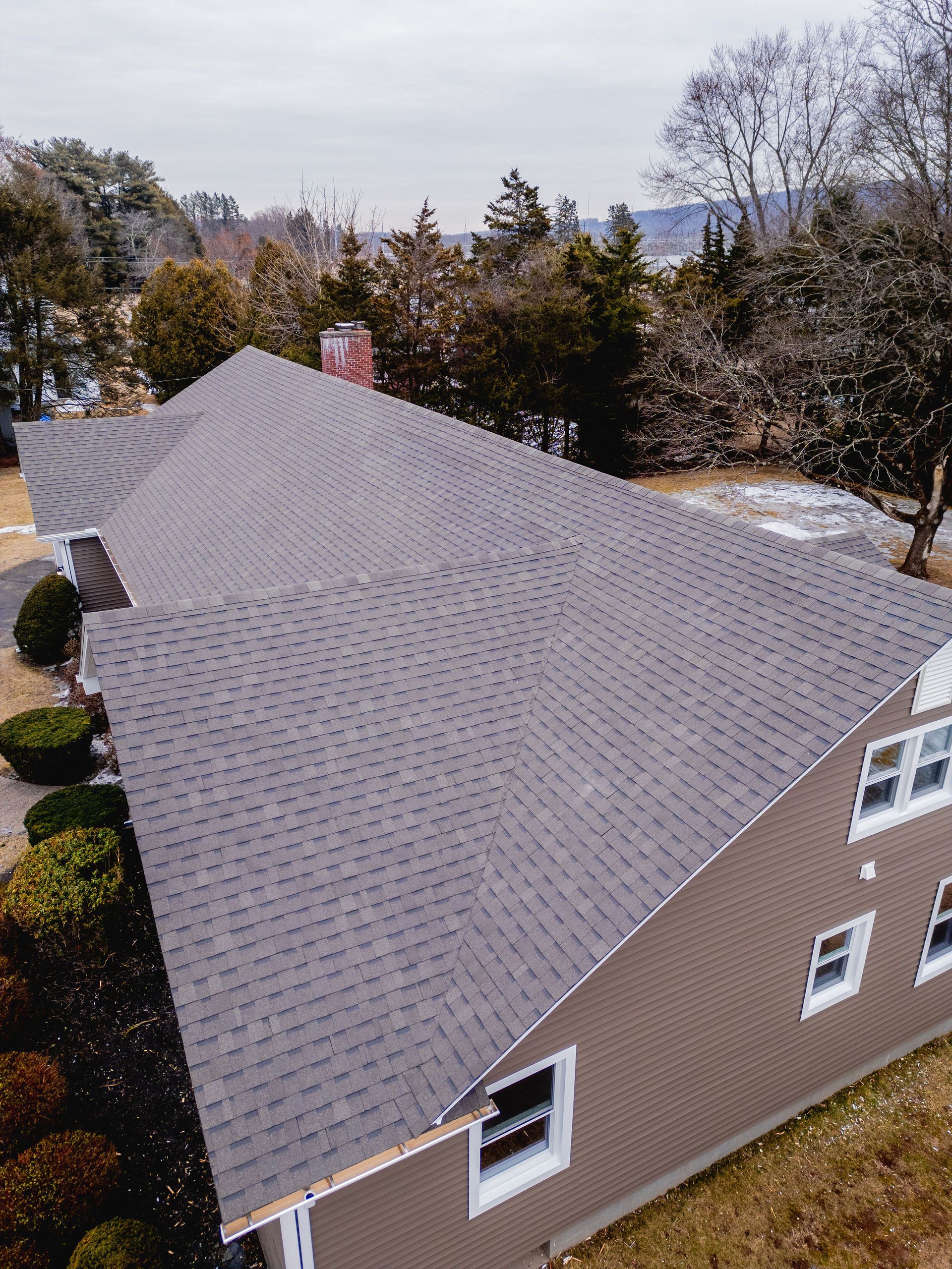 Gray roof on a brown house with white-trimmed windows; surrounded by trees and shrubs on a cloudy day.