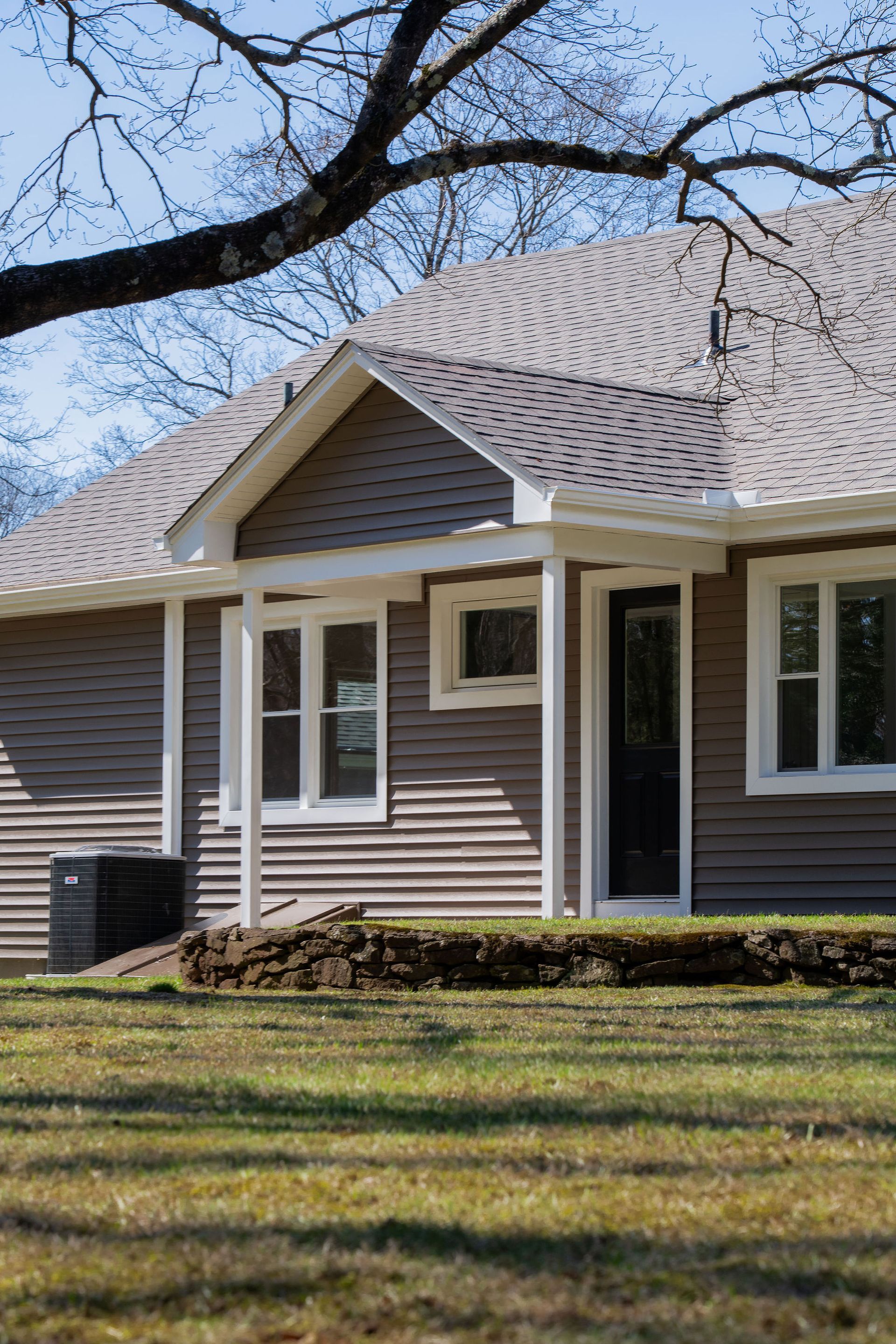 Tan house with white trim, dark door, and a porch under a tree in a yard.