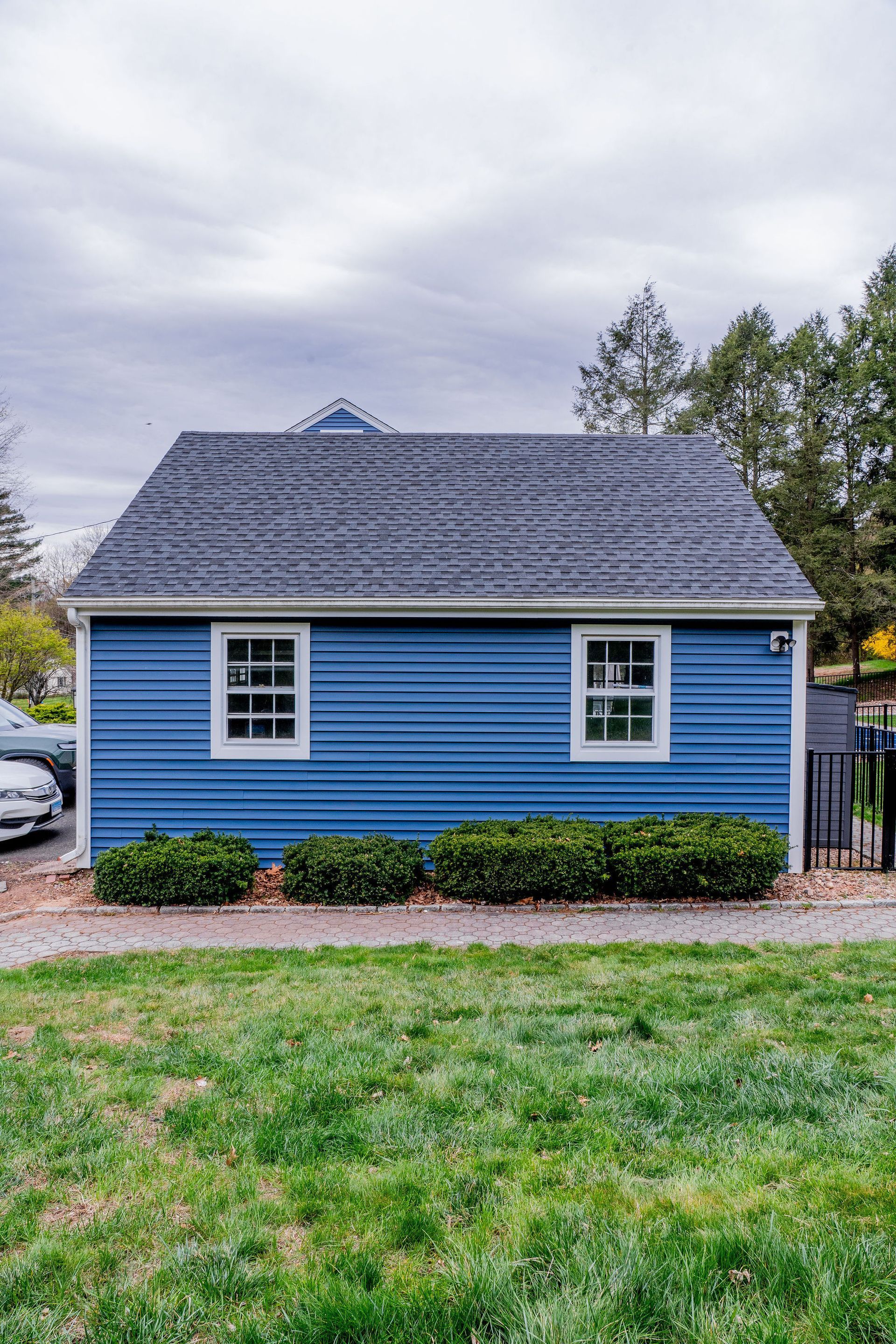 Blue-sided shed with white-framed windows and dark roof, framed by greenery and lawn, under a cloudy sky.
