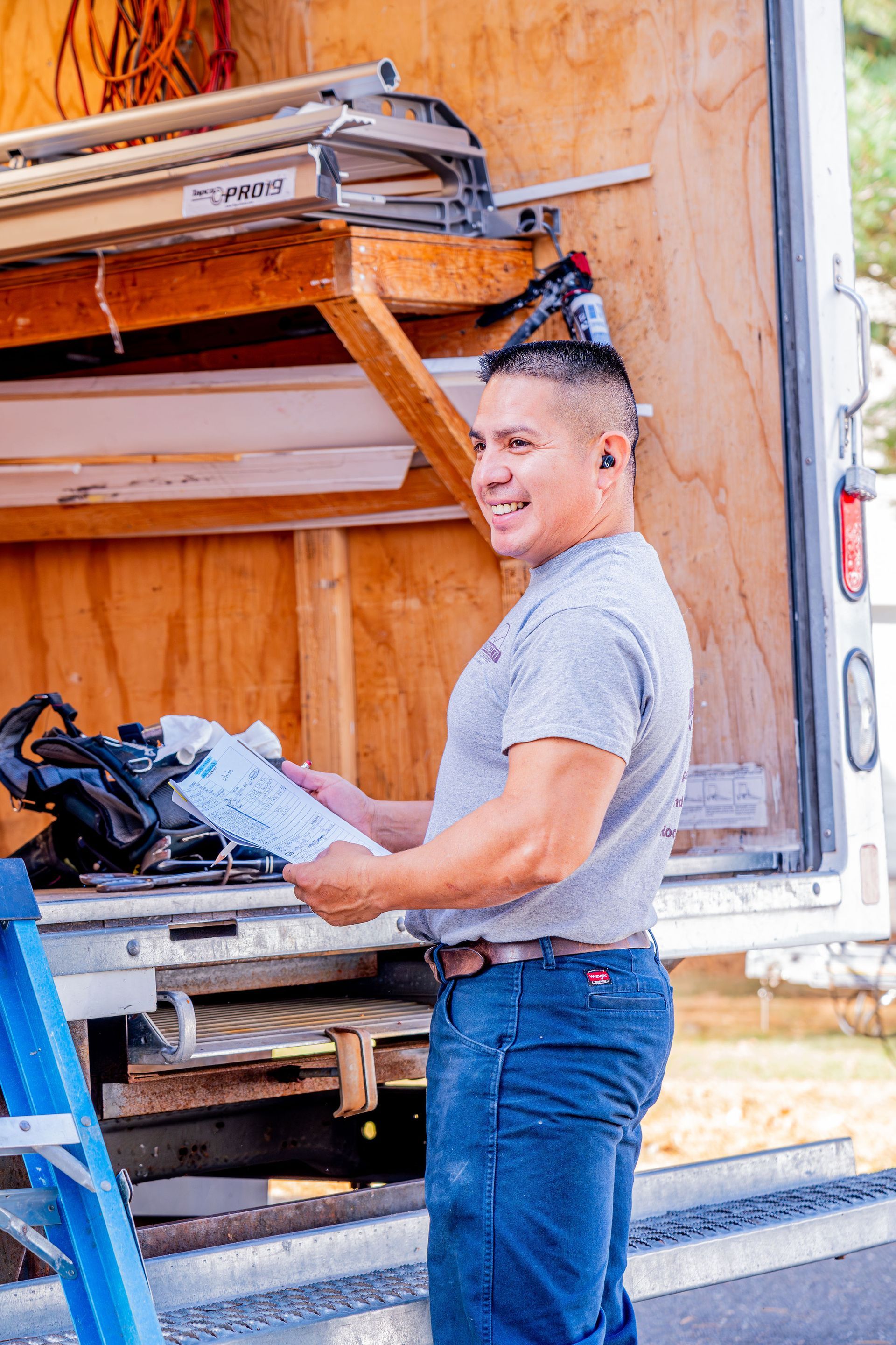 Man standing by a truck holding papers, smiling. He wears work clothes and ear protection.