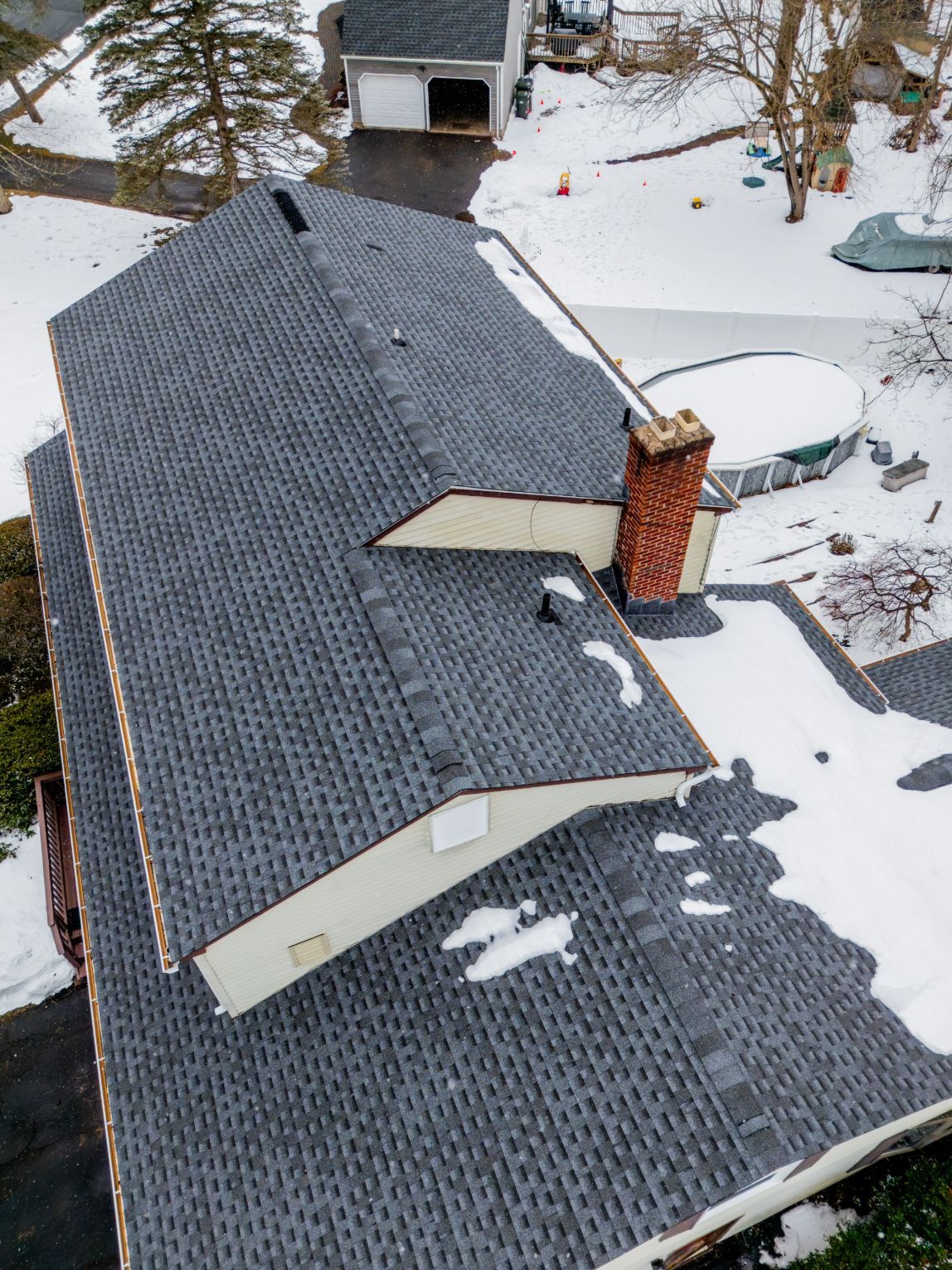 Snow-covered house roof with dark gray shingles and a red brick chimney.