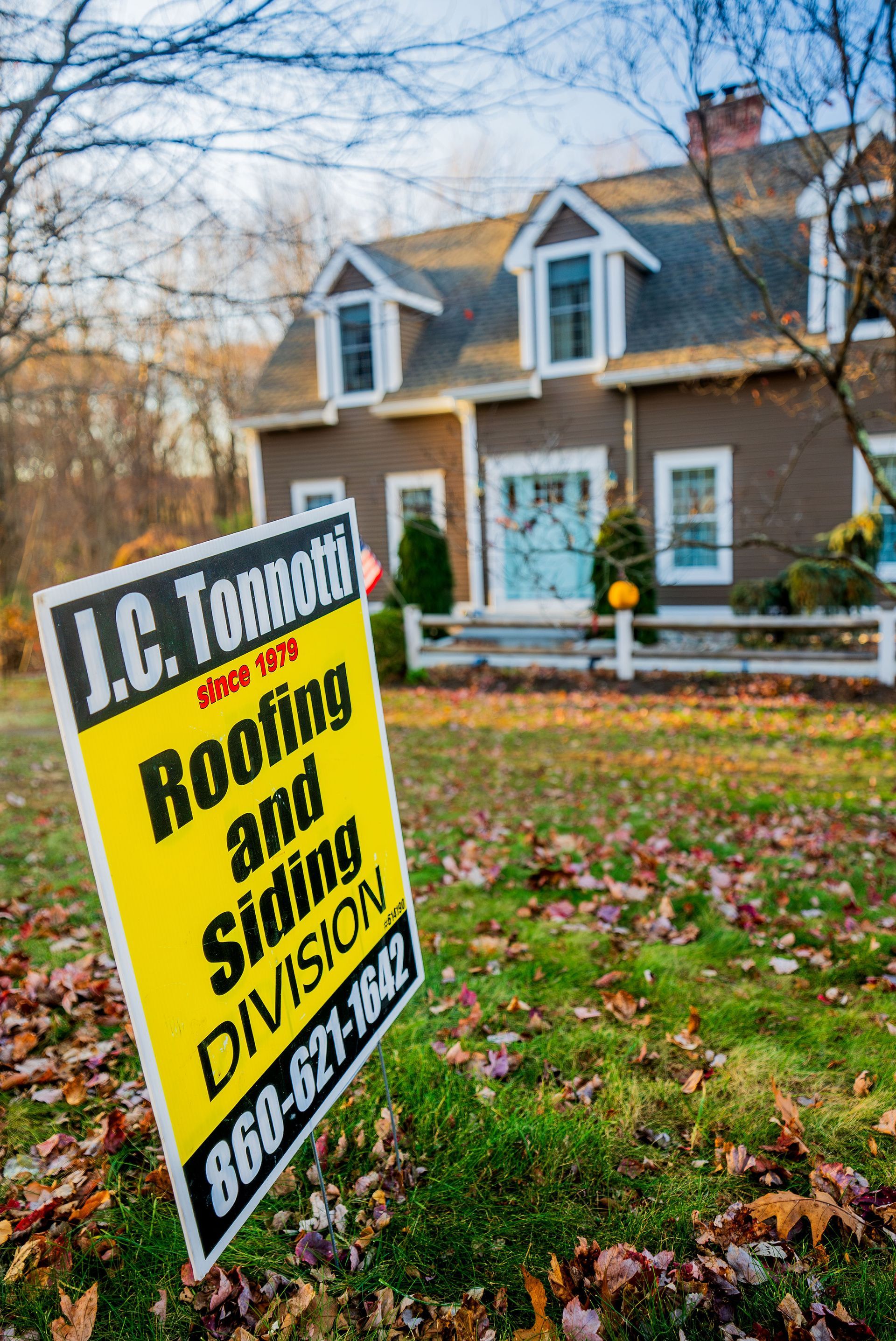 Yellow sign for roofing company in front of a brown house with dormers; foliage and autumn leaves.